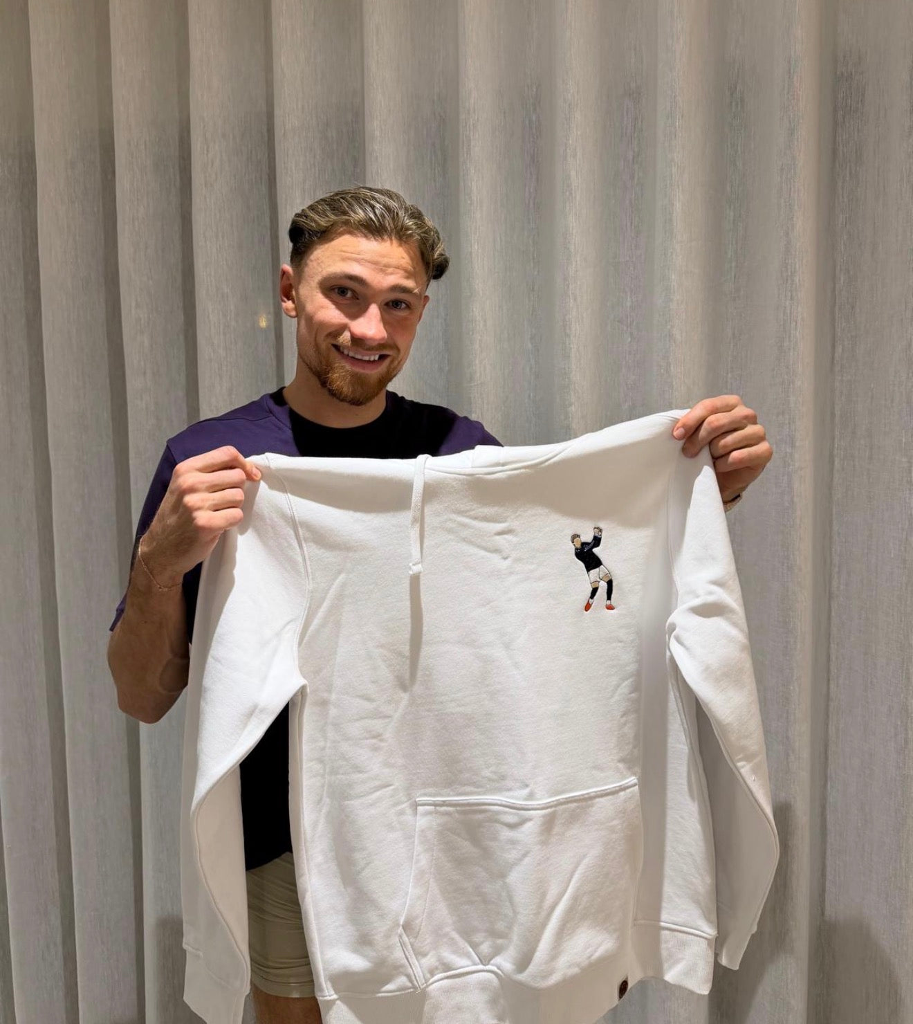 Matty Cash holding a custom white hoodie with a small graphic of his iconic golf celebration.